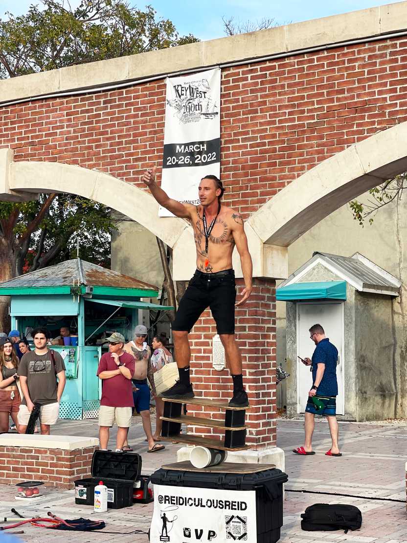 A street performer balancing on a wooden board on top of a cylinder. The man is shirtless with chest tattoos