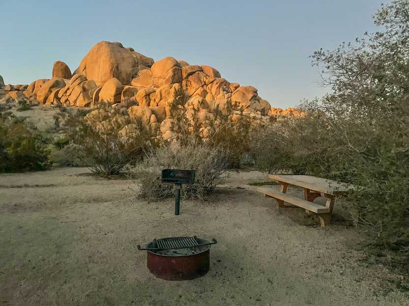 A campsite with a picnic table, fire pit and grill. Not far behind the campsite is a giant piles of boulders.