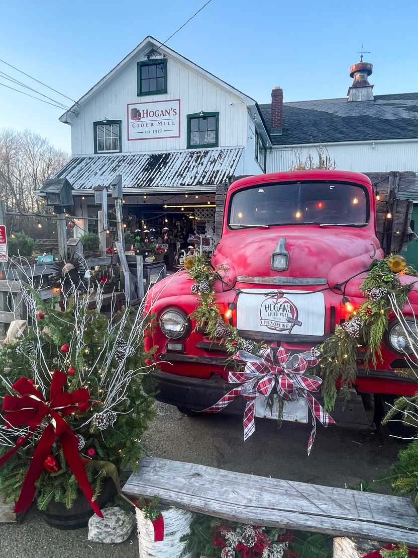 The exterior of Hogan's Cider Mill. There is a vintage red truck adorned with garland and a red bow. The general store in the background is white.