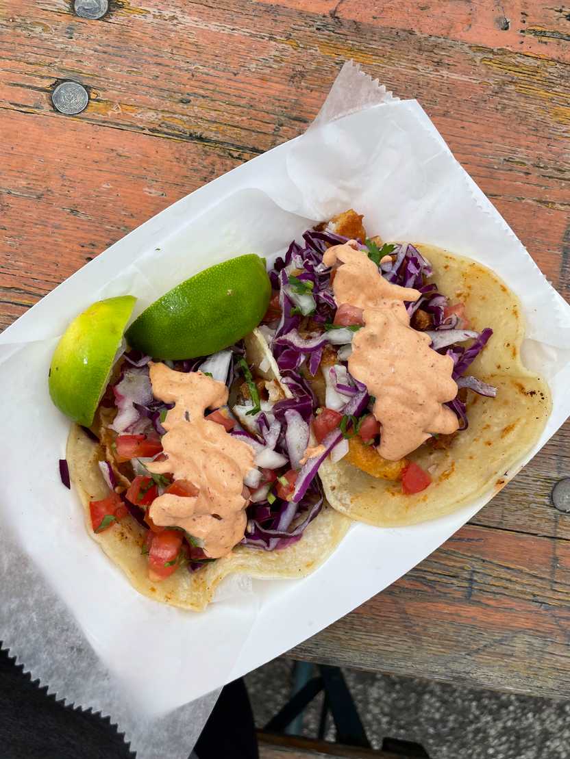 Two tacos from Miguel's Tacos. There is purple lettuce, chipotle sauce, tomatoes and lime wedges.