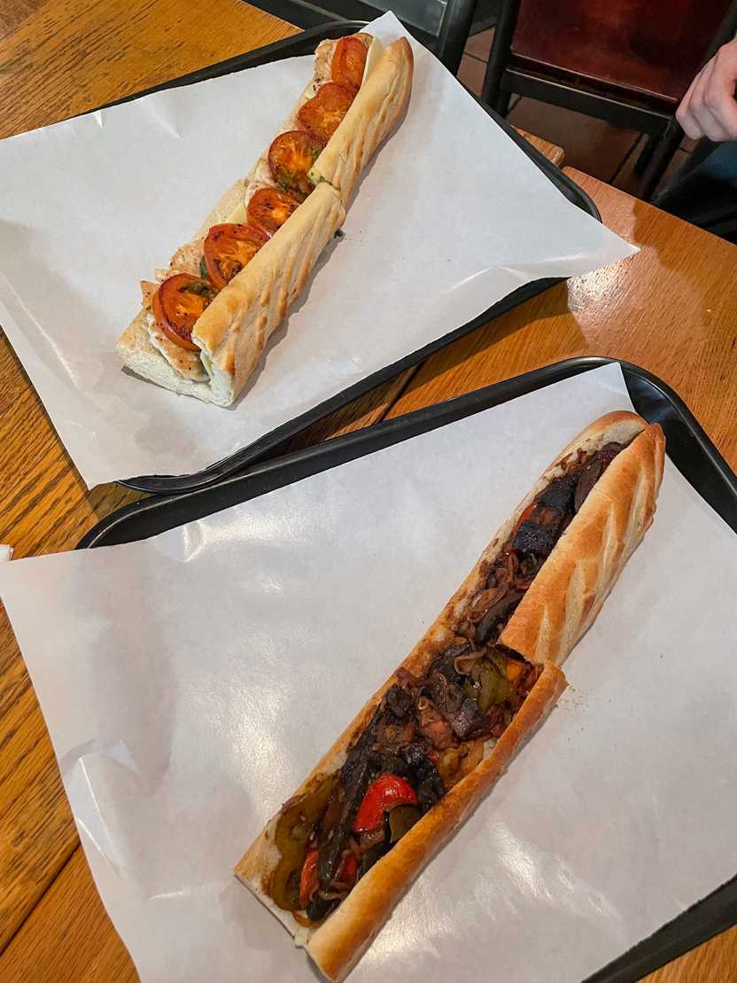 Two large subs from Dino's Cafe. One has veggies and the other has chicken and tomatoes.