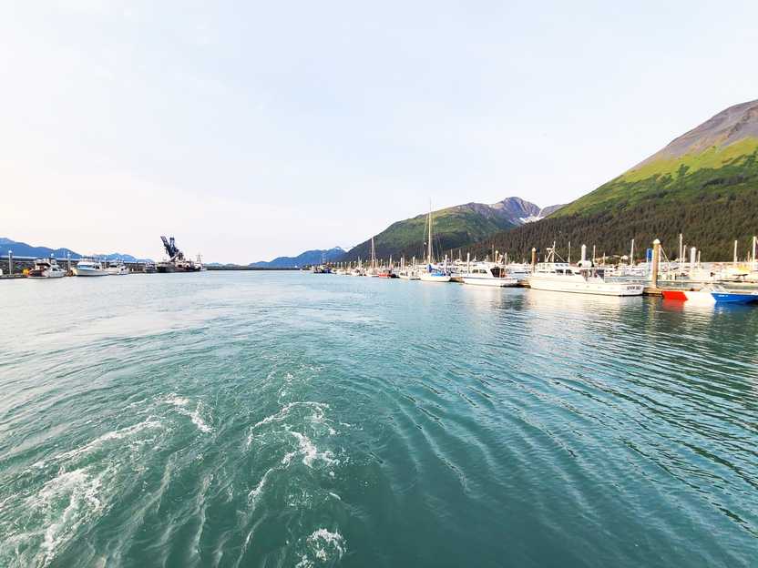 A bay with blue water, some boats along the shore and mountains in the background.