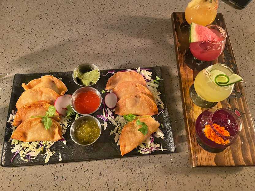 A plate of 8 mini tacos and tree dips in the center. Plus, there is a flight of 4 different margaritas in small glasses.