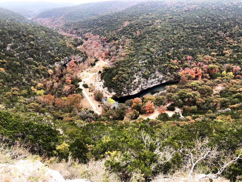 Looking down at a river next to a rock wall. Green trees cover the hills with bits of red foliage intermixed.
