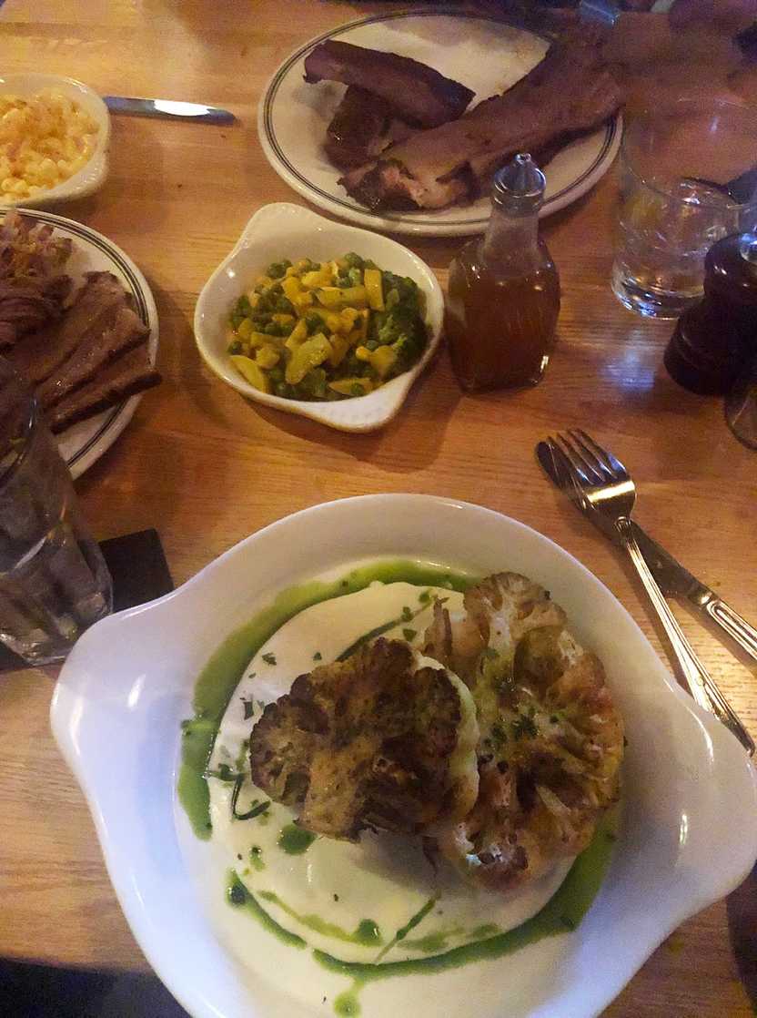 A plate of cauliflower steak, a BBQ veggies side and a plate of meat across the table.